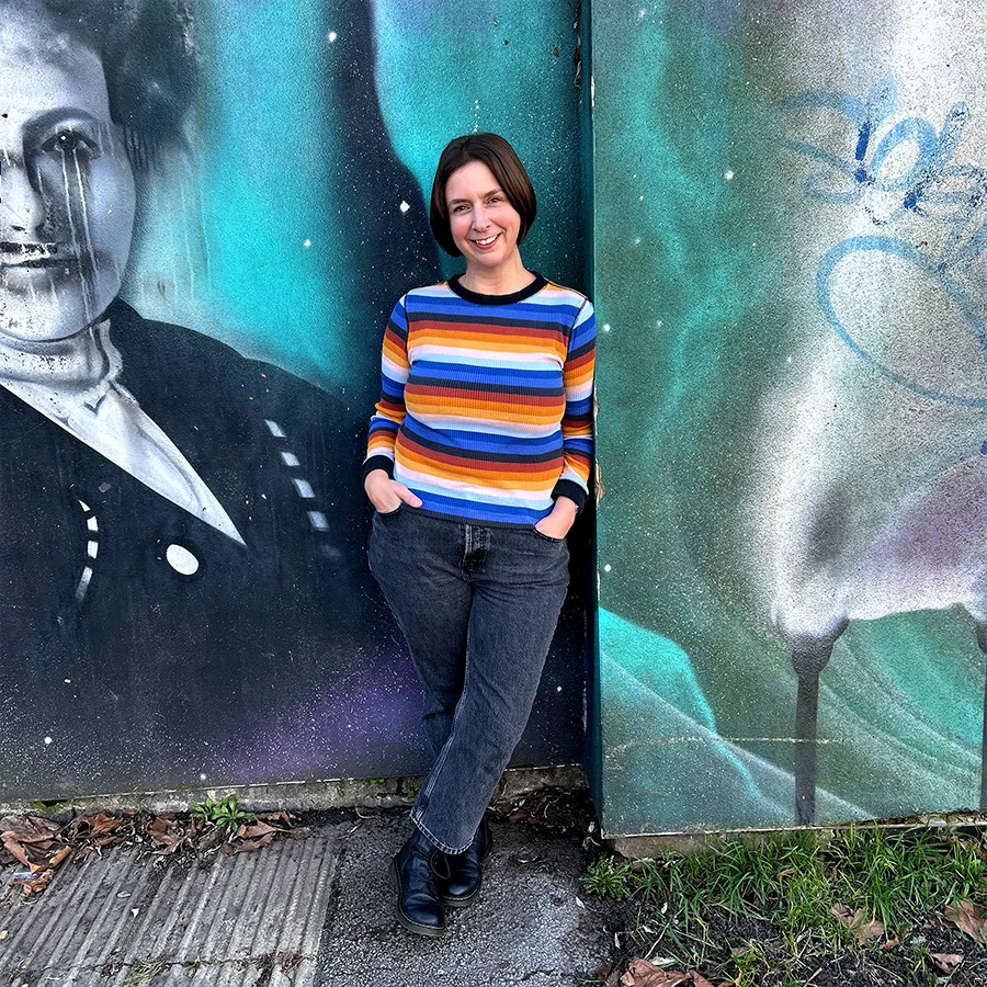Lou smiling in a bright, stripey top with a graffiti wall in the background.